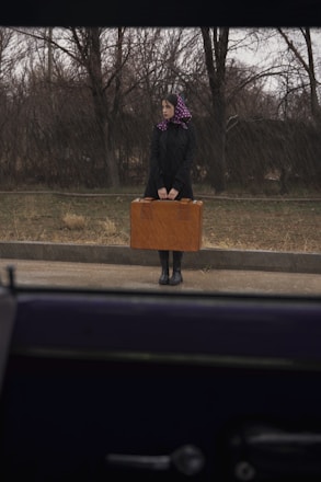 A woman stands on the roadside near barren trees, holding a large brown suitcase. She wears a black coat, boots, and a purple headscarf with white polka dots. The scene is observed from inside a vehicle.