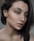 A close-up portrait of a person with short dark hair, minimal makeup, and a neutral facial expression. The lighting highlights the contours and details of the face.
