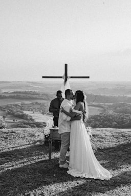A couple is embracing and kissing in front of a large wooden cross set against a scenic landscape. They appear to be partaking in a wedding ceremony on a grassy hilltop. Behind the couple, an officiant is present, dressed in dark clothing. The background features rolling hills and a vast open sky, captured in black and white.