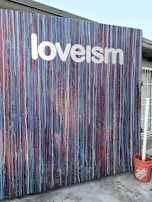 The image features an abstract art piece on a vertical surface adorned with drips and streaks of various colors. The word 'loveism' is written in white across the top portion. There is a red bucket placed near the bottom right corner, and part of a building structure is visible in the background.