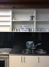 A minimalist kitchen scene with open white shelves displaying a coffee maker, various mugs, glasses, and a lightbox sign with the text 'WE COULD HAVE HAD IT ALL.' Below, a countertop with a stainless steel sink, a dish rack, and a cloth draped over the sink area is visible. The overall atmosphere is tidy and understated.