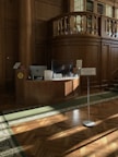 The scene depicts a classic, wood-paneled interior with a small reception desk adorned with a computer monitor, papers, and COVID-19 protective measures like a transparent barrier. Above the desk, there is a mezzanine-like structure with decorative columns and shelves filled with books. Sunlight streams through windows, casting patterns on the wooden floor and green carpet.