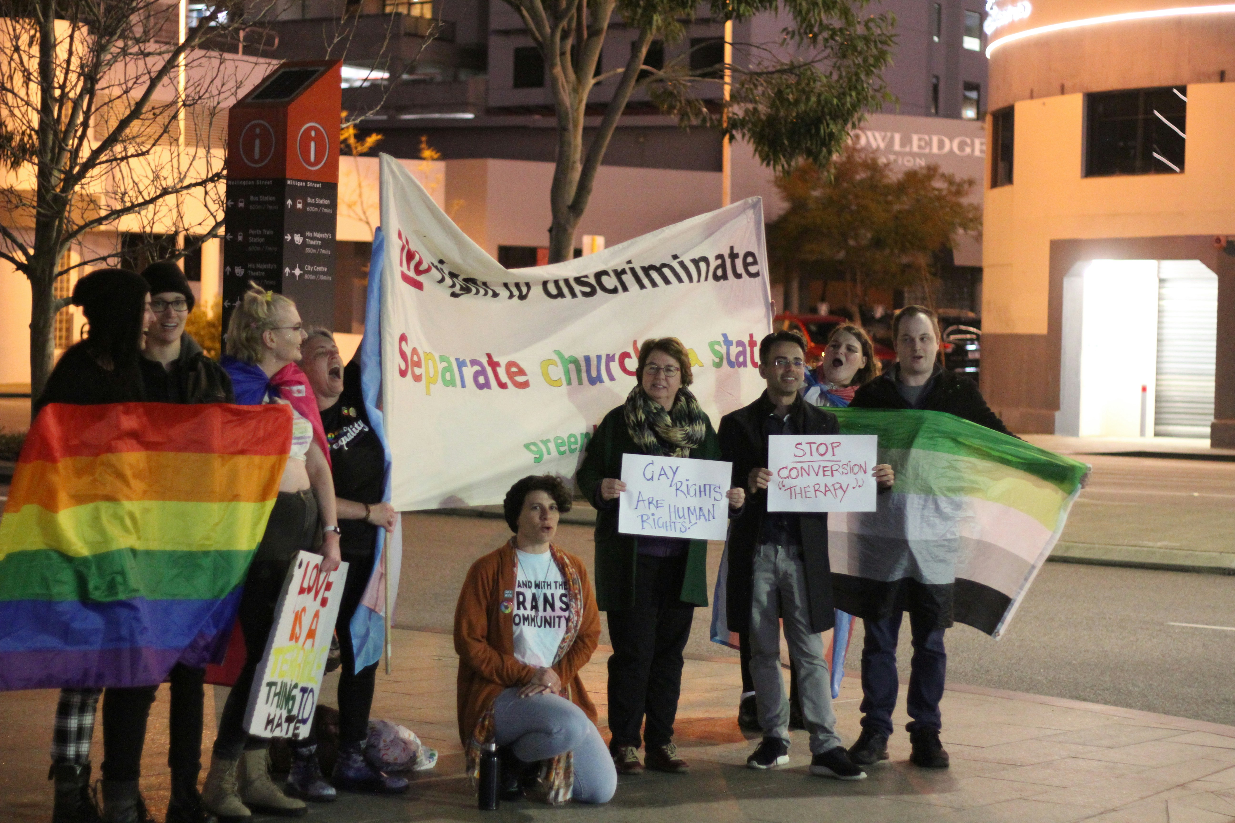 A diverse group of transgender individuals standing together with serious expressions, holding placards related to rights and inclusion.