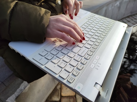 Hands with red nail polish typing on a white laptop keyboard, set outdoors, partially reflecting the surroundings. The person is wearing a green jacket.
