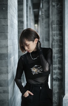 A person stands between brick pillars, wearing a black sheer top over dark clothing. Their pose conveys introspection, with their head slightly bowed and eyes closed. The somber setting is accentuated by the grayscale bricks.