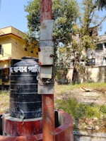 A large black water tank sits atop a cylindrical base made of red bricks. Beside it, a tall red pole stands, attached with an electrical meter box. In the background, there are trees with green foliage and residential buildings with a yellow exterior. The ground is overgrown with grass and some scattered foliage.