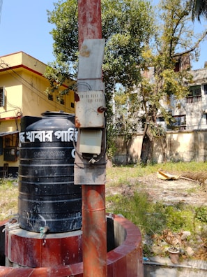 A large black water tank sits atop a cylindrical base made of red bricks. Beside it, a tall red pole stands, attached with an electrical meter box. In the background, there are trees with green foliage and residential buildings with a yellow exterior. The ground is overgrown with grass and some scattered foliage.