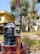 A large black water tank sits atop a cylindrical base made of red bricks. Beside it, a tall red pole stands, attached with an electrical meter box. In the background, there are trees with green foliage and residential buildings with a yellow exterior. The ground is overgrown with grass and some scattered foliage.