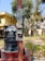 A large black water tank sits atop a cylindrical base made of red bricks. Beside it, a tall red pole stands, attached with an electrical meter box. In the background, there are trees with green foliage and residential buildings with a yellow exterior. The ground is overgrown with grass and some scattered foliage.