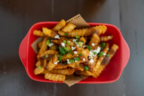 Loaded fries overflowing with cheese and sauce in a black rounded container, highlighted with golden-orange accents.