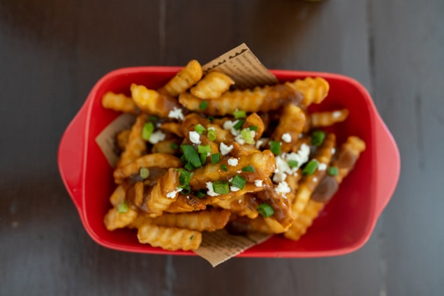 A red dish filled with crispy crinkle-cut fries topped with a savory sauce, garnished with chopped green onions and crumbled cheese, resting on a piece of decorative paper.