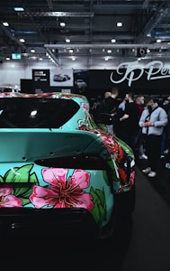 A car brightly decorated with floral and graphic patterns is parked indoors at an exhibition. The vibrant design features large pink flowers and intricate leaf motifs against a teal background. Several people are gathered around, indicating a public event or car show.
