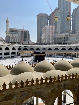 A view of the Kaaba at the center of the Grand Mosque in Mecca, surrounded by people performing the Tawaf ritual. The mosque's distinctive domed architecture, arches, and intricate design elements are visible in the foreground. In the background, towering skyscrapers and cranes are part of the cityscape.