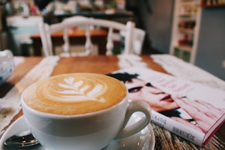A cappuccino with a beautifully crafted latte art sits on a wooden table next to a book. The background suggests a cozy cafe setting with blurred chairs and shelving.