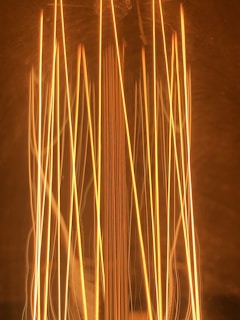Vertical filaments of a glowing light bulb create an intricate pattern with warm tones and a soft, ambient glow. The filaments are arranged in parallel and slightly crisscross, emanating a rich, golden hue.