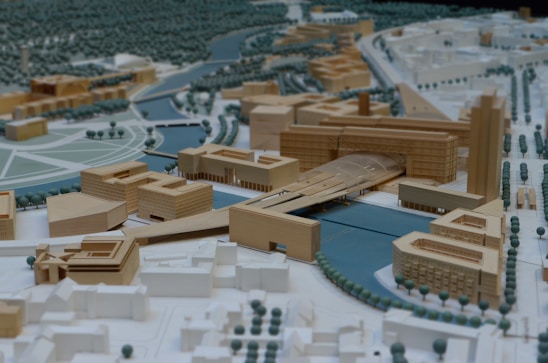 A detailed architectural scale model of an urban area featuring a variety of buildings arranged around a central waterway. The structures are made from wood and have a mix of geometric shapes. There are simulated roads and pathways with evenly spaced trees, some arranged in circular patterns. The model includes a prominent bridge structure crossing the water and connecting different parts of the area.