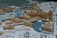 A detailed architectural scale model of an urban area featuring a variety of buildings arranged around a central waterway. The structures are made from wood and have a mix of geometric shapes. There are simulated roads and pathways with evenly spaced trees, some arranged in circular patterns. The model includes a prominent bridge structure crossing the water and connecting different parts of the area.