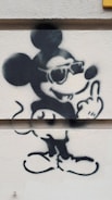 A graffiti-style drawing on a wall depicts a well-known cartoon mouse character wearing sunglasses and giving a cheeky expression with one hand gesturing. The artwork uses bold black outlines on a light-colored surface.