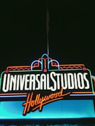 Bright neon signage displays the words 'Universal Studios Hollywood' against a dark backdrop. The lettering is illuminated in white and orange, with decorative accents in blue, red, and green neon tubes.