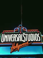 Bright neon signage displays the words 'Universal Studios Hollywood' against a dark backdrop. The lettering is illuminated in white and orange, with decorative accents in blue, red, and green neon tubes.