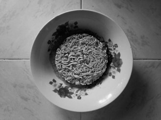 A package of instant noodles placed on a decorative plate with floral patterns. The setting appears to be on a tiled surface, and the image is in black and white.