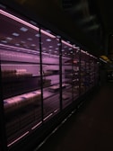 A dimly lit supermarket aisle featuring a long row of refrigerated shelves with glass doors. The pinkish-purple lighting casts a moody glow, highlighting packaged items like eggs and other grocery products. The shelves appear partially stocked.