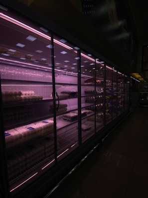 A dimly lit supermarket aisle featuring a long row of refrigerated shelves with glass doors. The pinkish-purple lighting casts a moody glow, highlighting packaged items like eggs and other grocery products. The shelves appear partially stocked.
