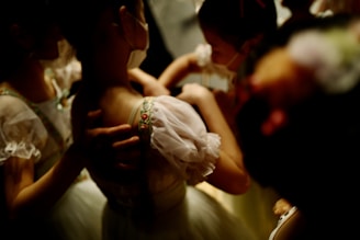 A group of children wearing light-colored costumes with delicate lace and floral patterns. The lighting is dim, casting a warm glow on their outfits. They appear to be preparing for a performance, possibly adjusting each other's garments with focused and gentle expressions.