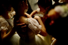 A group of children wearing light-colored costumes with delicate lace and floral patterns. The lighting is dim, casting a warm glow on their outfits. They appear to be preparing for a performance, possibly adjusting each other's garments with focused and gentle expressions.