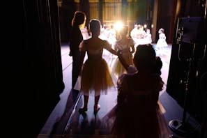A group of young ballerinas dressed in white tutus waits in the wings of a theater stage. Warm stage lighting casts long shadows and silhouettes. Some ballerinas are holding hands, and an adult figure stands nearby. On stage, more ballerinas are performing under bright lights.