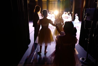 A group of young ballerinas dressed in white tutus waits in the wings of a theater stage. Warm stage lighting casts long shadows and silhouettes. Some ballerinas are holding hands, and an adult figure stands nearby. On stage, more ballerinas are performing under bright lights.