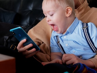 A young child with an expression of surprise or excitement is seated on someone's lap, gazing at a smartphone held by an adult. The child is wearing a light blue shirt with dark suspenders, and the background suggests they are sitting on a dark-colored couch.