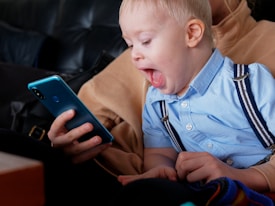 A young child with an expression of surprise or excitement is seated on someone's lap, gazing at a smartphone held by an adult. The child is wearing a light blue shirt with dark suspenders, and the background suggests they are sitting on a dark-colored couch.