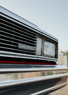 Close-up view of the front grille of a Chevrolet vehicle, featuring the brand emblem prominently displayed. The image captures the headlight and part of the bumper with reflections visible on the metallic surface. The sky in the background is clear and subtly blurred.