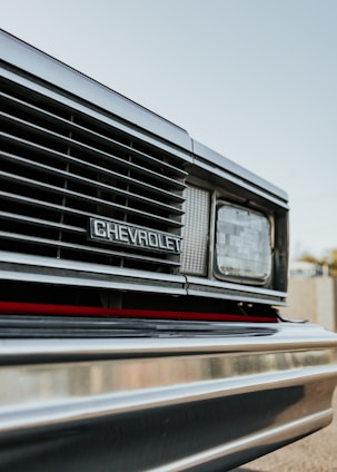 Close-up view of the front grille of a Chevrolet vehicle, featuring the brand emblem prominently displayed. The image captures the headlight and part of the bumper with reflections visible on the metallic surface. The sky in the background is clear and subtly blurred.