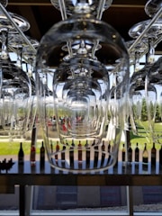 Upside-down wine glasses are neatly arranged in rows, creating a symmetrical and reflective pattern. Behind the glasses, an outdoor scene with lush greenery and a line of wine bottles is visible through large windows.