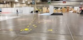 A large convention center hall arranged for an event setup with scattered boxes, crates, and equipment. There are partitions covered with white drapes, directional signs hanging from the ceiling, and yellow taped lines on the floor. A person is walking through the hall, and equipment is positioned sporadically.