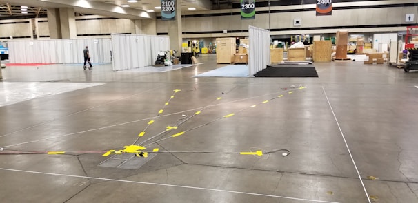 A large convention center hall arranged for an event setup with scattered boxes, crates, and equipment. There are partitions covered with white drapes, directional signs hanging from the ceiling, and yellow taped lines on the floor. A person is walking through the hall, and equipment is positioned sporadically.