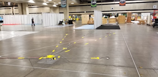 A large convention center hall arranged for an event setup with scattered boxes, crates, and equipment. There are partitions covered with white drapes, directional signs hanging from the ceiling, and yellow taped lines on the floor. A person is walking through the hall, and equipment is positioned sporadically.