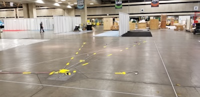 A large convention center hall arranged for an event setup with scattered boxes, crates, and equipment. There are partitions covered with white drapes, directional signs hanging from the ceiling, and yellow taped lines on the floor. A person is walking through the hall, and equipment is positioned sporadically.