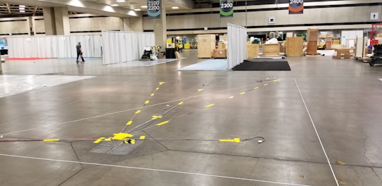 A large convention center hall arranged for an event setup with scattered boxes, crates, and equipment. There are partitions covered with white drapes, directional signs hanging from the ceiling, and yellow taped lines on the floor. A person is walking through the hall, and equipment is positioned sporadically.