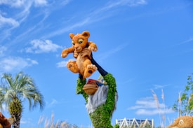 A colorful sculpture depicting a baboon holding a lion cub high in the air, made from greenery and plant materials. The sky is clear with a few clouds. There are palm trees and ornamental grasses visible in the background.
