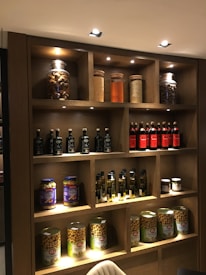 A wooden shelf filled with various jars, bottles, and cans of food items. The top shelf contains large jars with ingredients like nuts and grains. The second shelf features bottles of olive oil and red vinegar. Below, jars of pickles and larger cans of preserved goods are displayed.