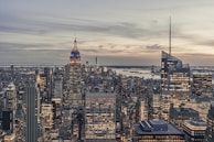 A cityscape featuring numerous skyscrapers with the prominent Empire State Building lit in blue and white. The skyline is under a cloudy sky, with a partial view of a river in the background. The buildings are densely packed, showing a vibrant, urban environment.