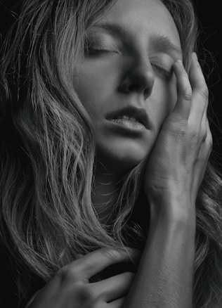 A black and white photograph of a person with closed eyes, gently touching their face with their hand. Their expression conveys a sense of serenity or introspection.