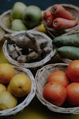 Several wicker baskets contain a variety of fruits and vegetables, including green apples, oranges, lemons, carrots, ginger, and cucumbers. The arrangement is on a mustard yellow background, and the lighting adds a rustic and natural aesthetic.