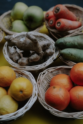 Several wicker baskets contain a variety of fruits and vegetables, including green apples, oranges, lemons, carrots, ginger, and cucumbers. The arrangement is on a mustard yellow background, and the lighting adds a rustic and natural aesthetic.