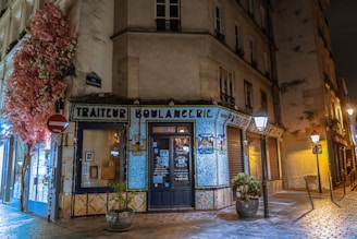 A charming street corner with a boulangerie decorated with mosaic tiles. The storefront is adorned with colorful floral arrangements and surrounded by warm street lighting. The building has an old-world European architecture style, and the cobblestone street adds to the quaint atmosphere.