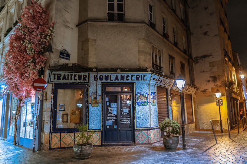 A charming street corner with a boulangerie decorated with mosaic tiles. The storefront is adorned with colorful floral arrangements and surrounded by warm street lighting. The building has an old-world European architecture style, and the cobblestone street adds to the quaint atmosphere.