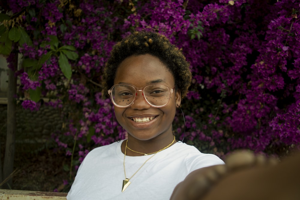 a woman with glasses smiling in front of purple flowers